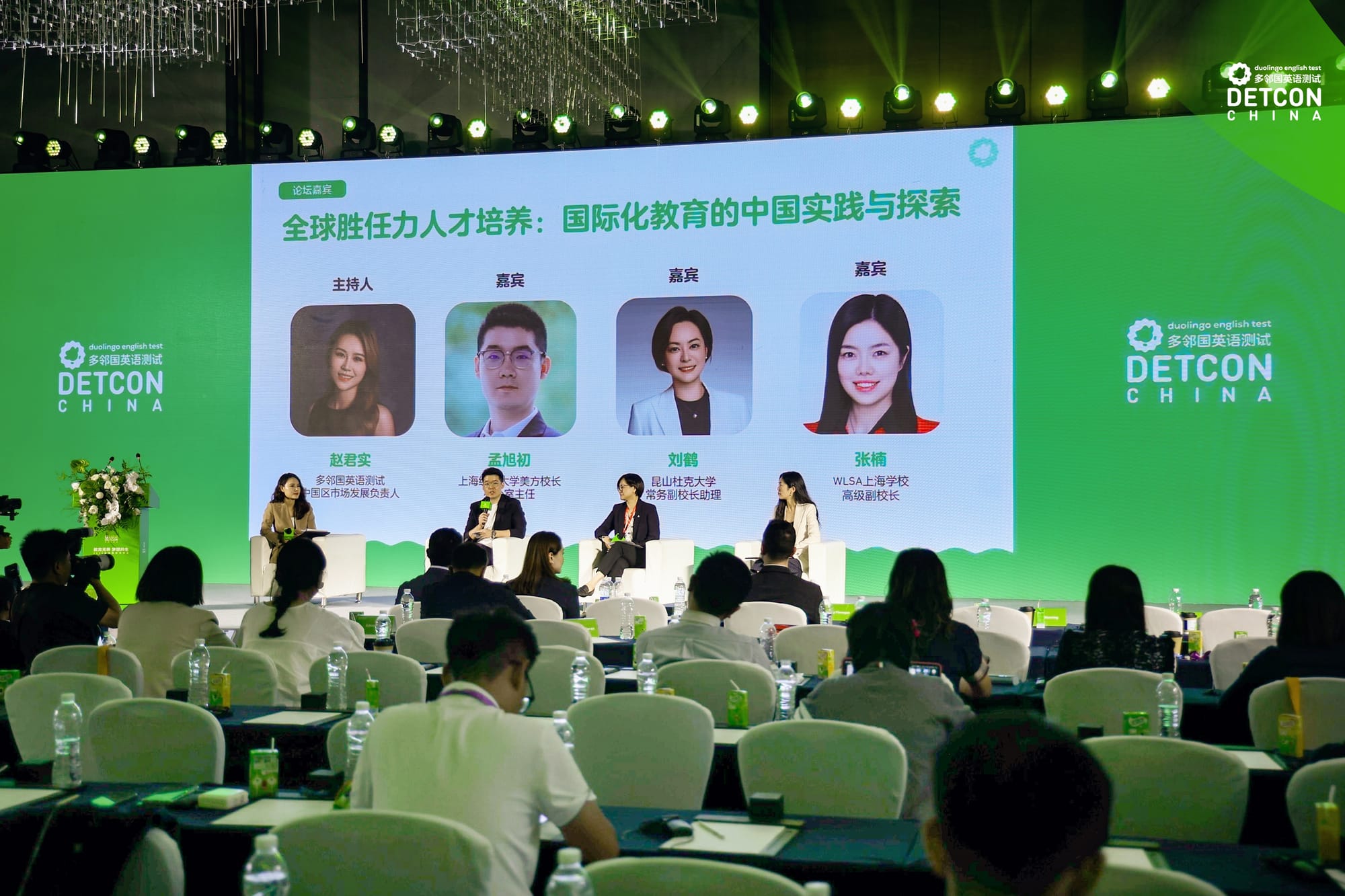 Four people sit on stage facing a crowd. Behind them, their headshots are displayed on screen. 