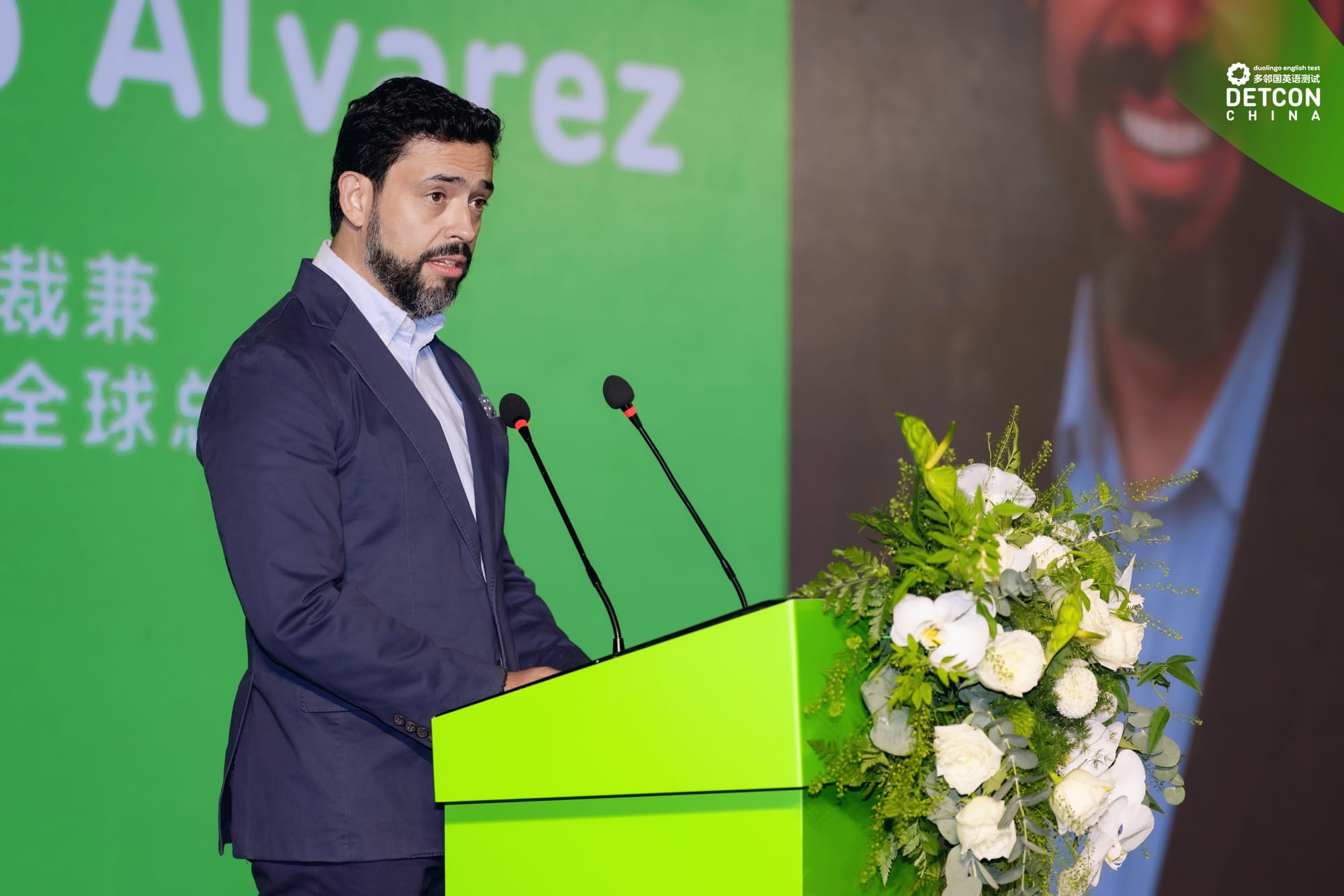 A man in a suit stands in front of a green podium, decorated with white flowers. He is speaking into a microphone. 