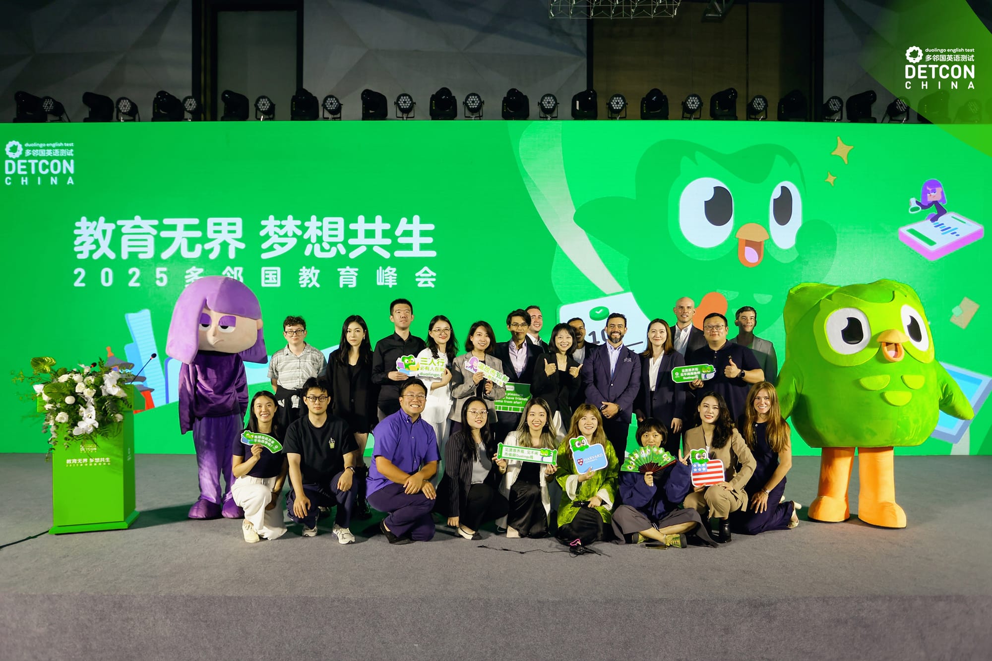 A group of people posing for a picture behind a green wall. On their right and left are people wearing Duolingo mascot suits of Duo and Lily.