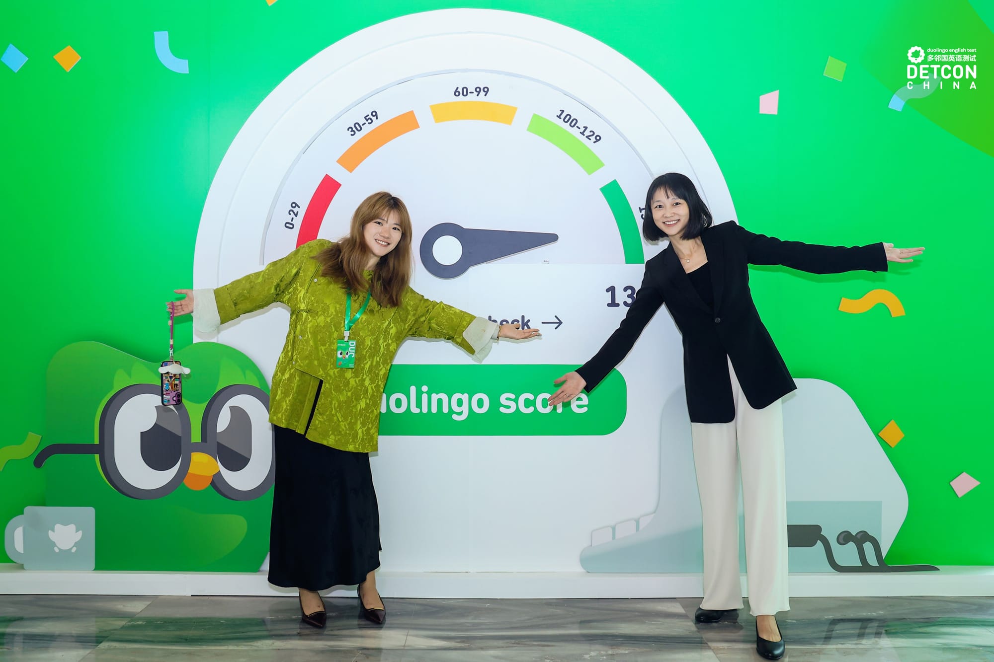 Two women stand with their arms outstretched in front of a green wall, which has a speedometer style graphic to track Duolingo score. 
