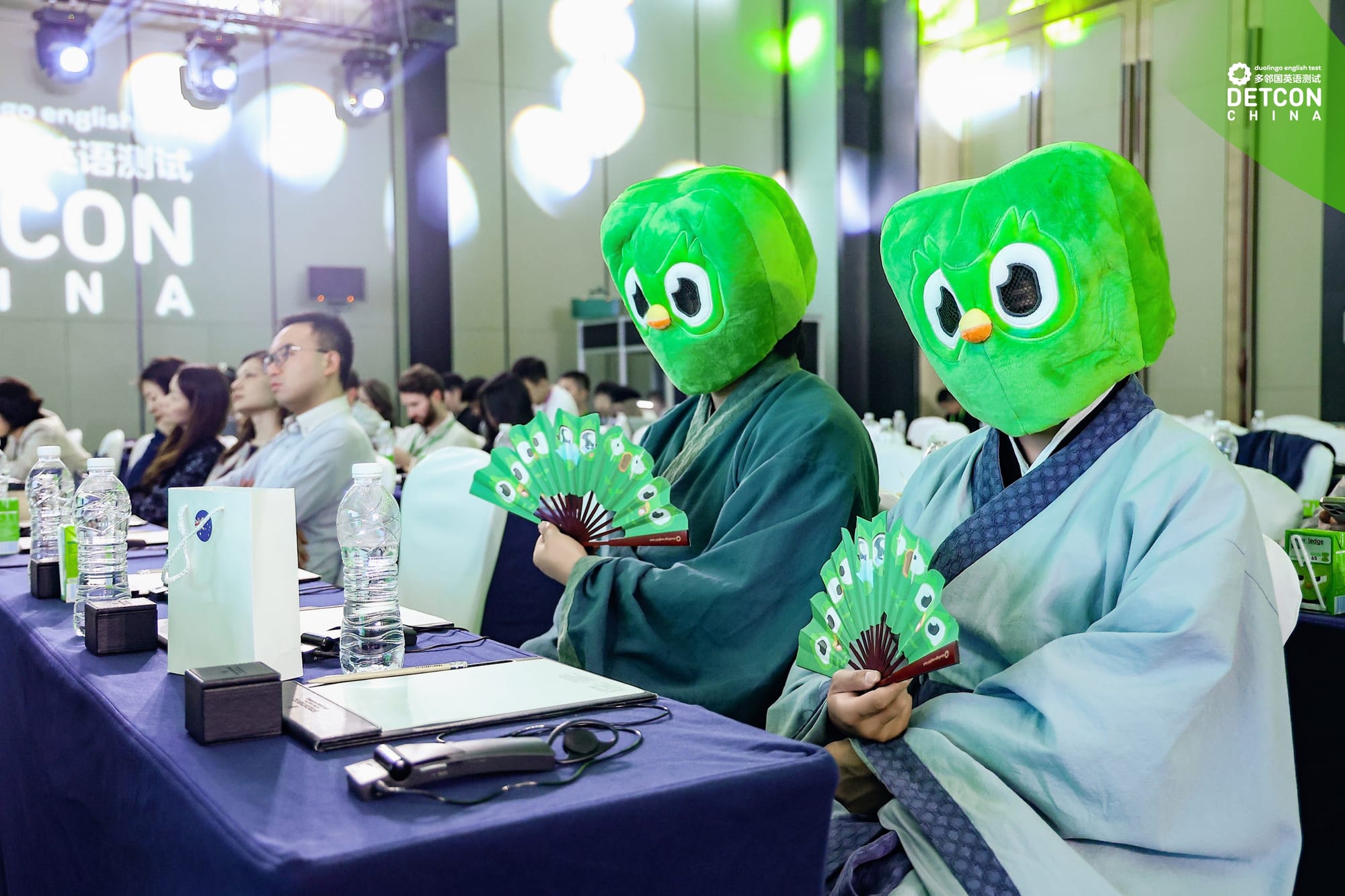Two people sit in an audience, wearing Green Duolingo masks and holding green Duolingo folding fans. 