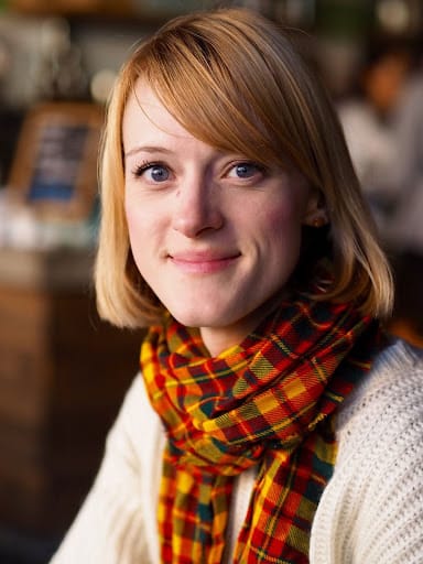 A headshot of a woman with short, strawberry blonde hair, wearing a red and orange plaid scarf and a white sweater