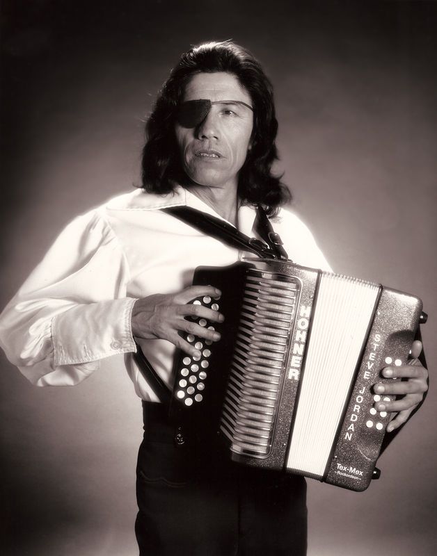An older Hispanic man, known as Esteban Jordan, with an eye patch and long hair in a white long-sleeve tunic holding an accordion.