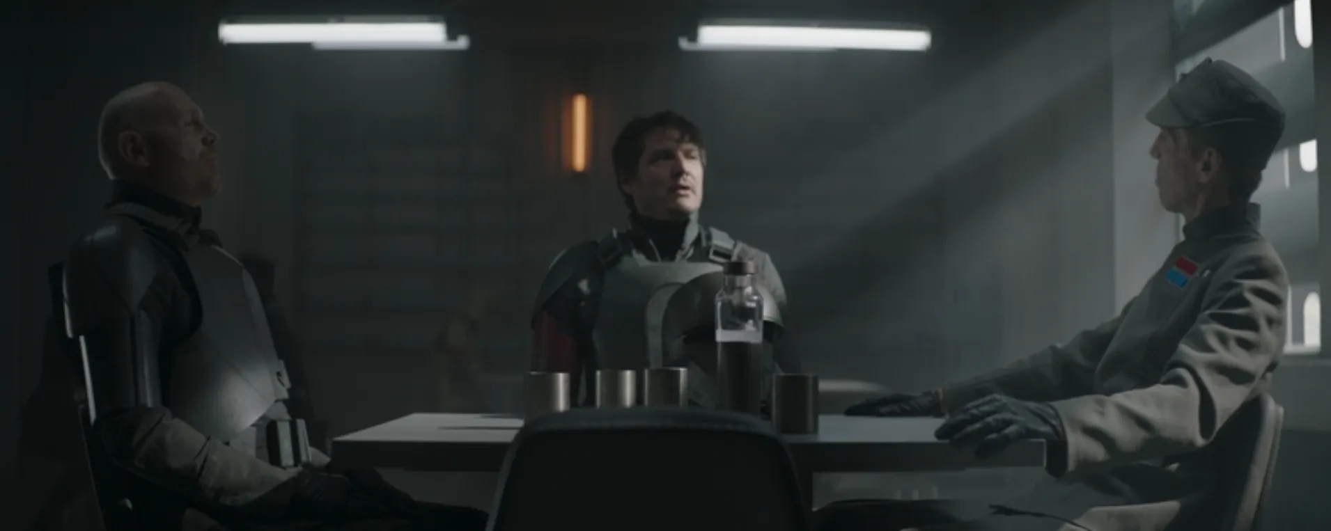 Din Djarin (Pedro Pascal), center, is seated at the center of the table facing the camera with Migs Mayfield (Bill Burr) on his right and an Imperial officer, Valin Hess (Richard Brake) on his left.