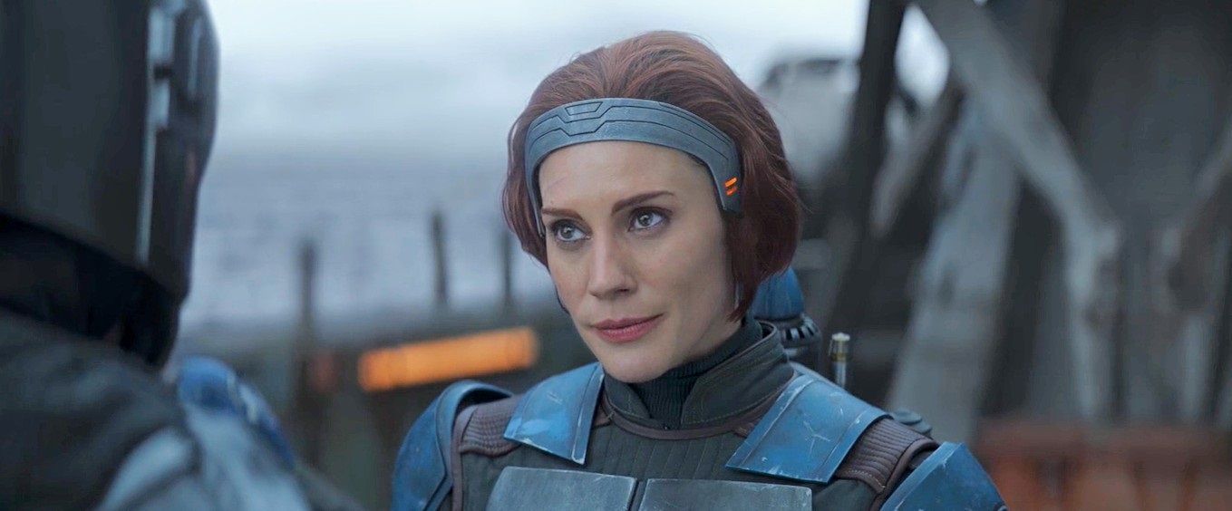 Bo-Katan Kryze (Katee Sackhoff) looks at Mando, who is visible on the corner of the screen. Her hair is red-brown and her armor is blue. They are on a ship with the sea visible behind them.
