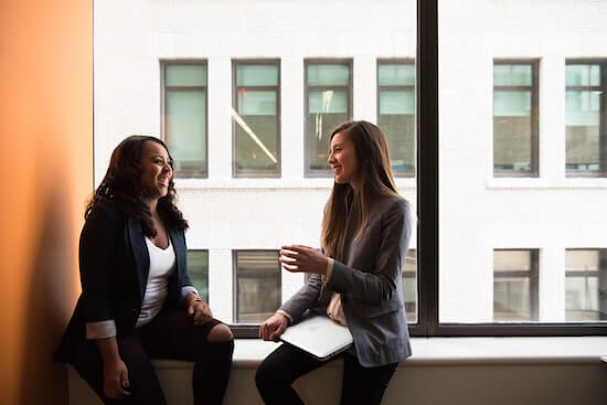 A photograph of two businesswomen sitting together and talking. They are both smiling.