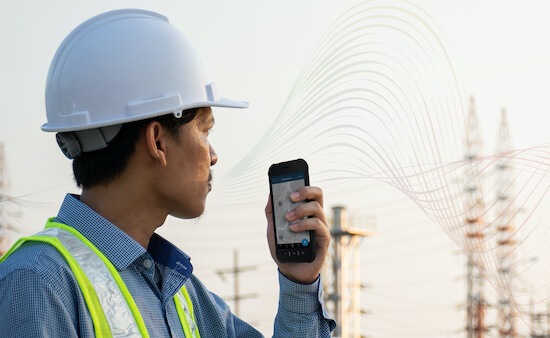 A man wearing a hard hat and safety vest is looking at some power lines while holding a mobile phone.