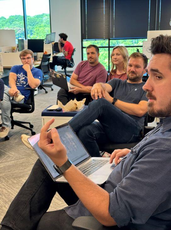 A photograph of the product squad in a regular meeting. Several people are seated in a circle in the middle of an office.