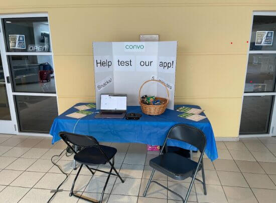 A photograph of the booth set-up. There's a table and two chairs, a basket full of snacks, a laptop computer, and a sign that says "Help test our app!"