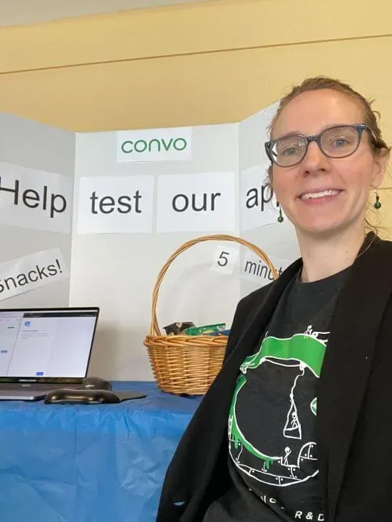 A photograph of Amanda standing in front of a booth. There's a sign visible on the booth that says, "Help test our app."
