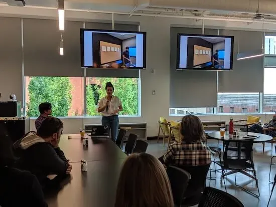 Nicole stands in front of a room of people holding a microphone and speaking.