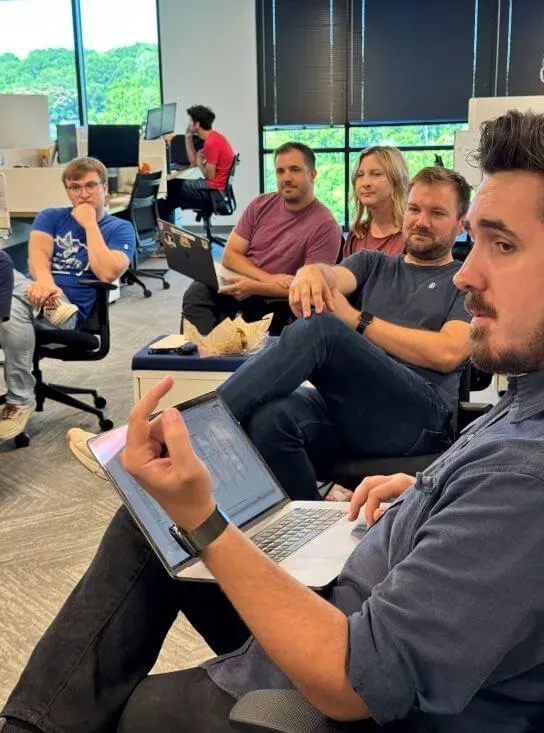 A photograph of the product squad in a regular meeting. Several people are seated in a circle in the middle of an office.