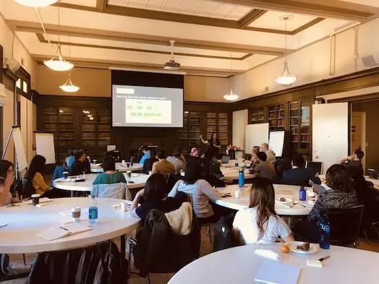 A large room with a workshop in session. Teresa is standing in the front of the room in front of a projector screen and groups of people are seated at tables.