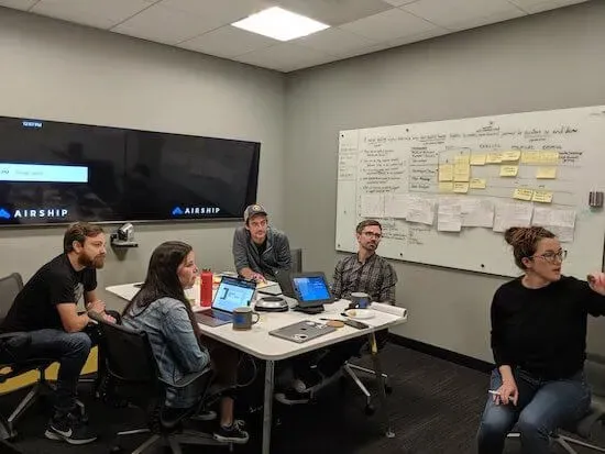 Lisa's team gathered around a table in a conference room. There's a whiteboard in the background covered in sticky notes.