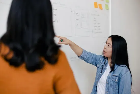 Two women standing in front of a white board with writing on it.