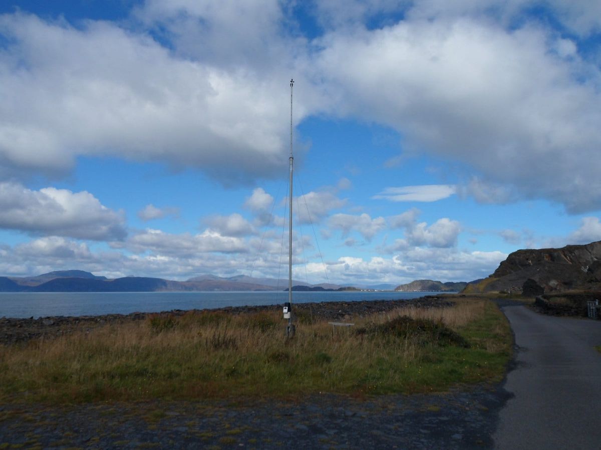 A tall thin mast with a small solar panel and monitoring unit stands in a grassy coastal field.