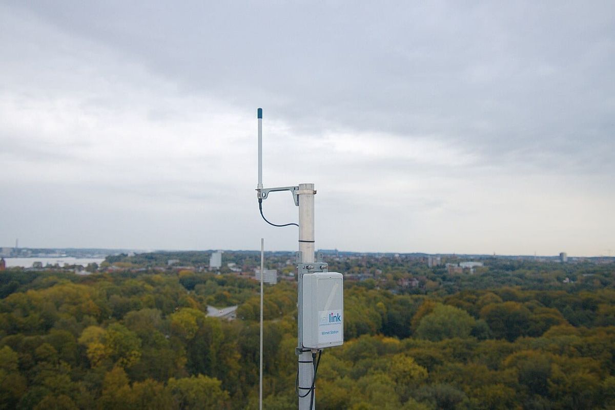 A Kerlink LoRaWAN gateway labeled 'Weenat Station' mounted on a metal pole overlooking a vast green landscape under cloudy skies.