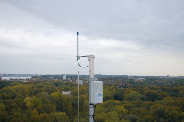 A Kerlink LoRaWAN gateway labeled 'Weenat Station' mounted on a metal pole overlooking a vast green landscape under cloudy skies.