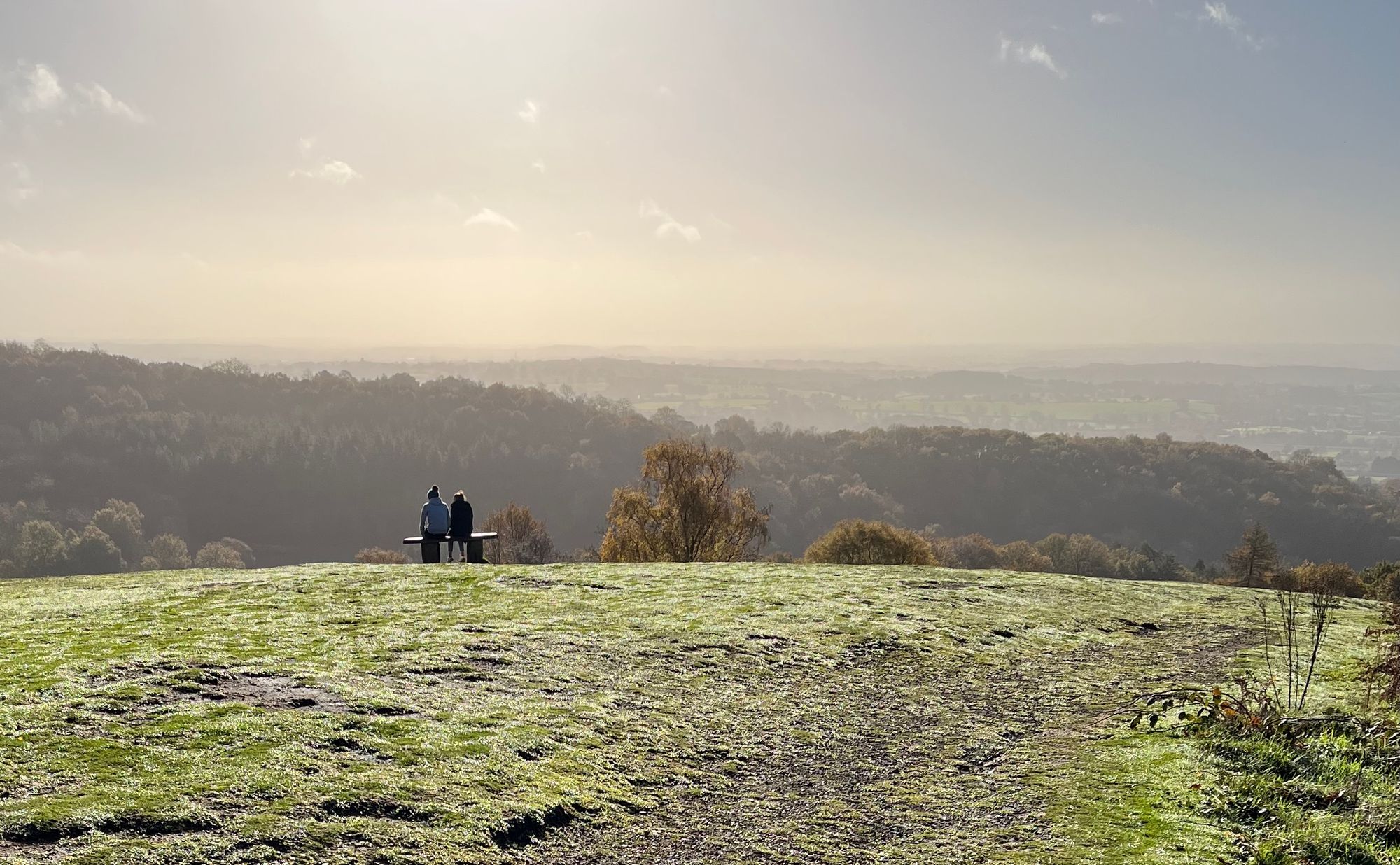 Two people on a bench at the top of a hill looking towards a sunny sky and trees and hills