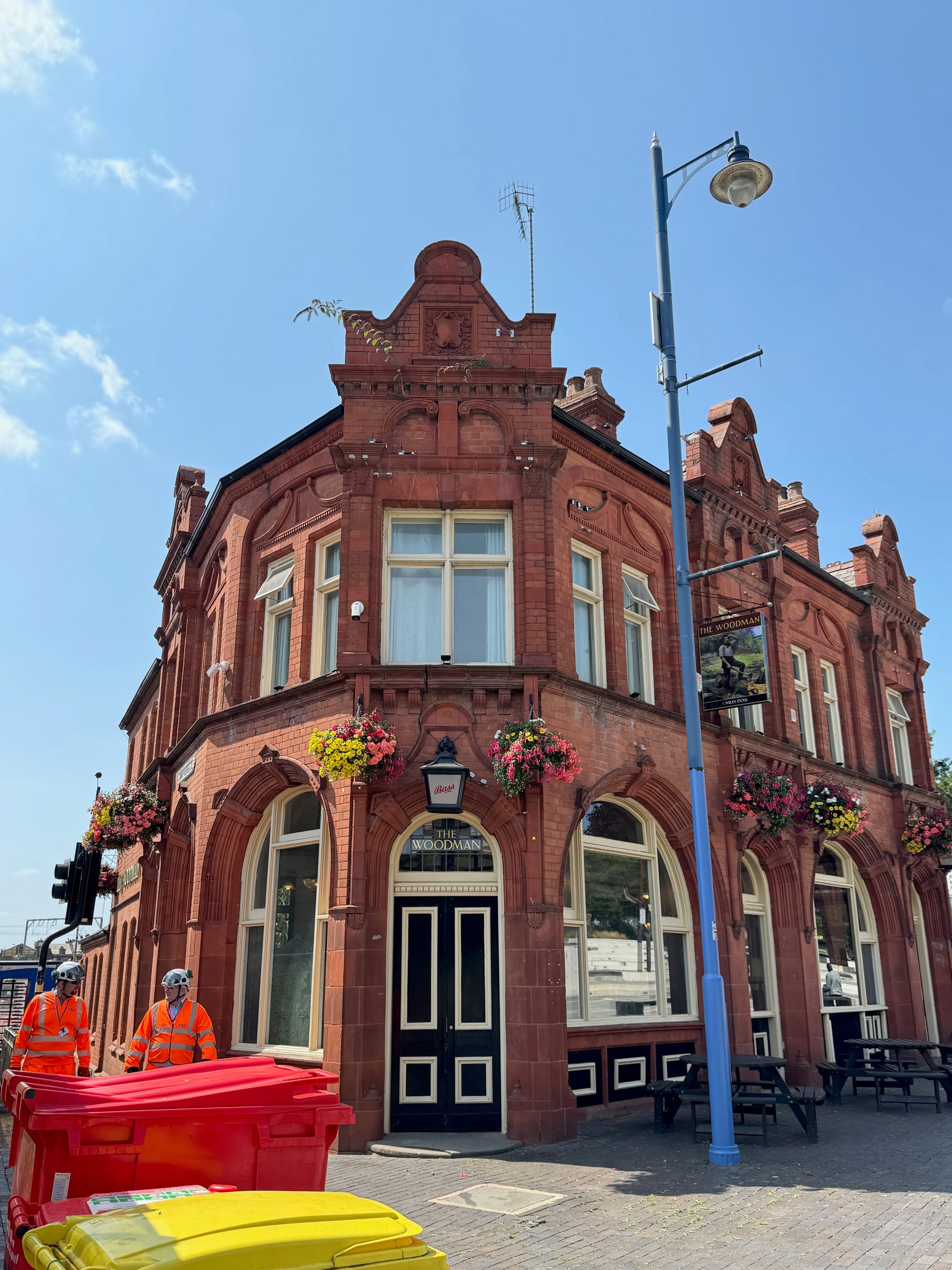 A red brick pub against a blue sky. Flowers in hanging baskets. Two workmen in PPE walk past