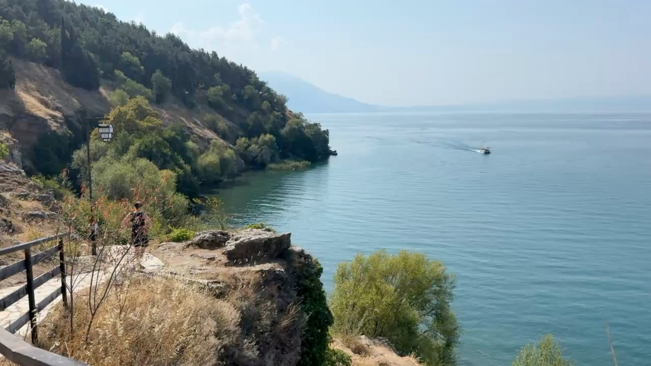 a woman on a pathway among shrubs by the side of a wide blue lake. a boat approaches. The sky is blue