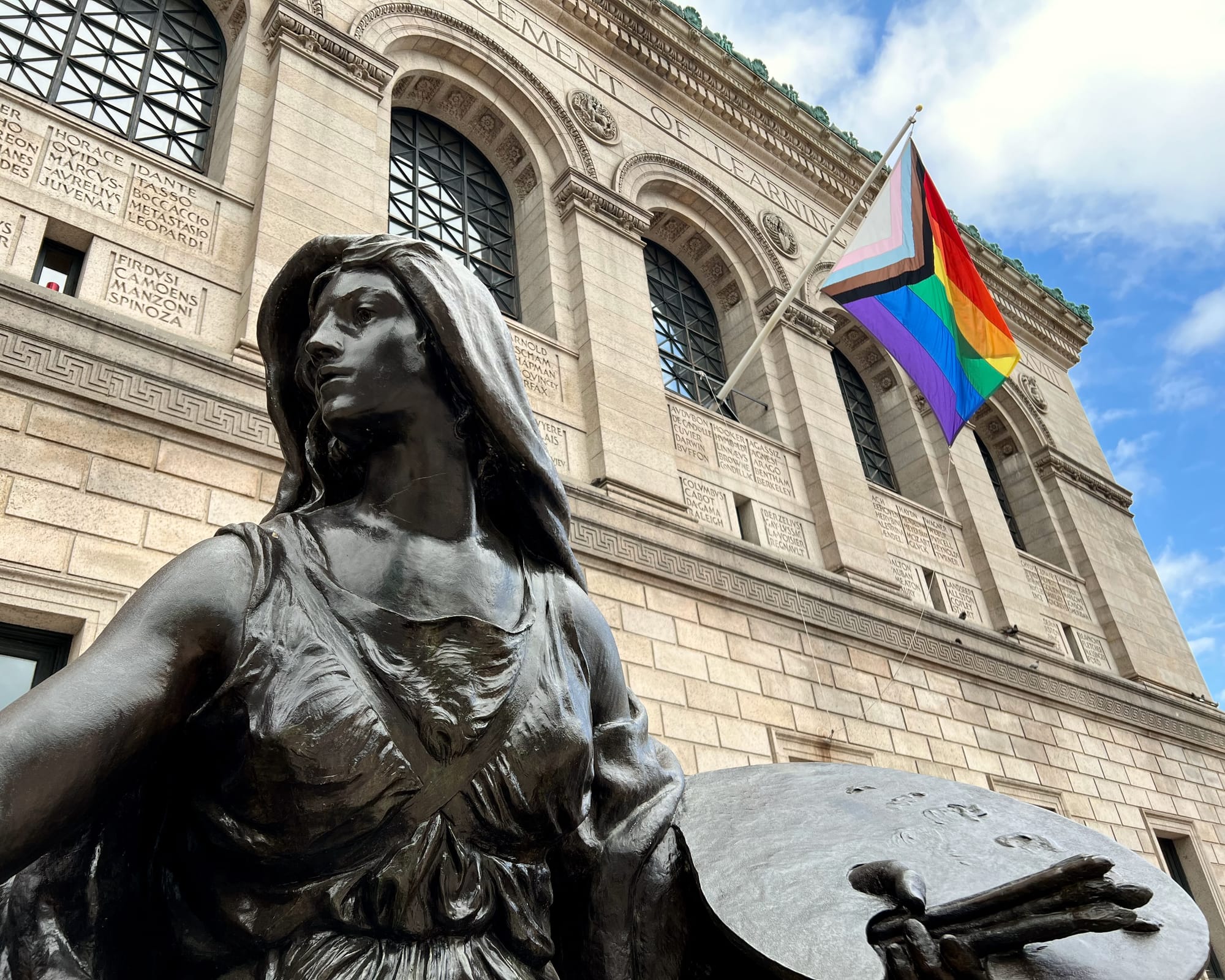 The outside of the Boston Public Library. A statue is in the foreground and a pride flag is hanging in the background.