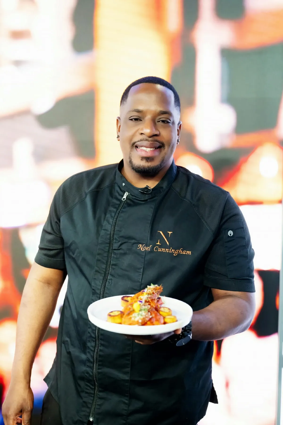 Chef Noel Cunningham in black chef whites, smiling and holding a plated Caribbean dish