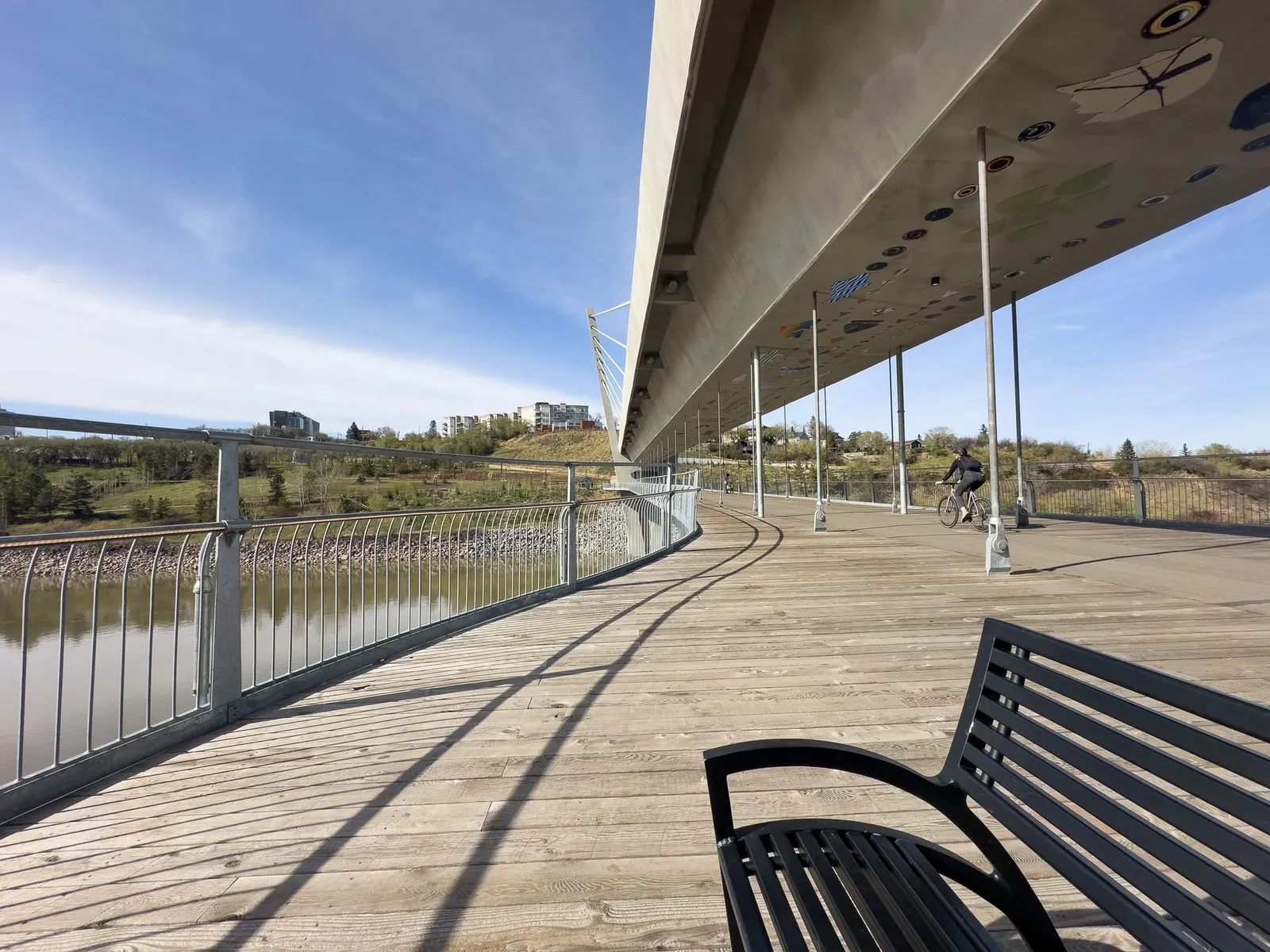 The riverside boardwalk curving beneath a modern bridge deck in Edmonton, a cyclist passing a bench