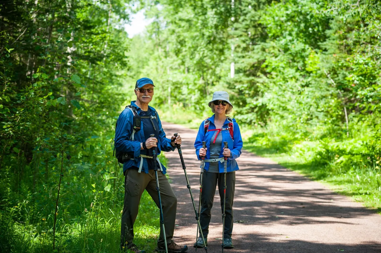 Two hikers with poles and blue jackets pausing on a tree-lined crushed-stone trail