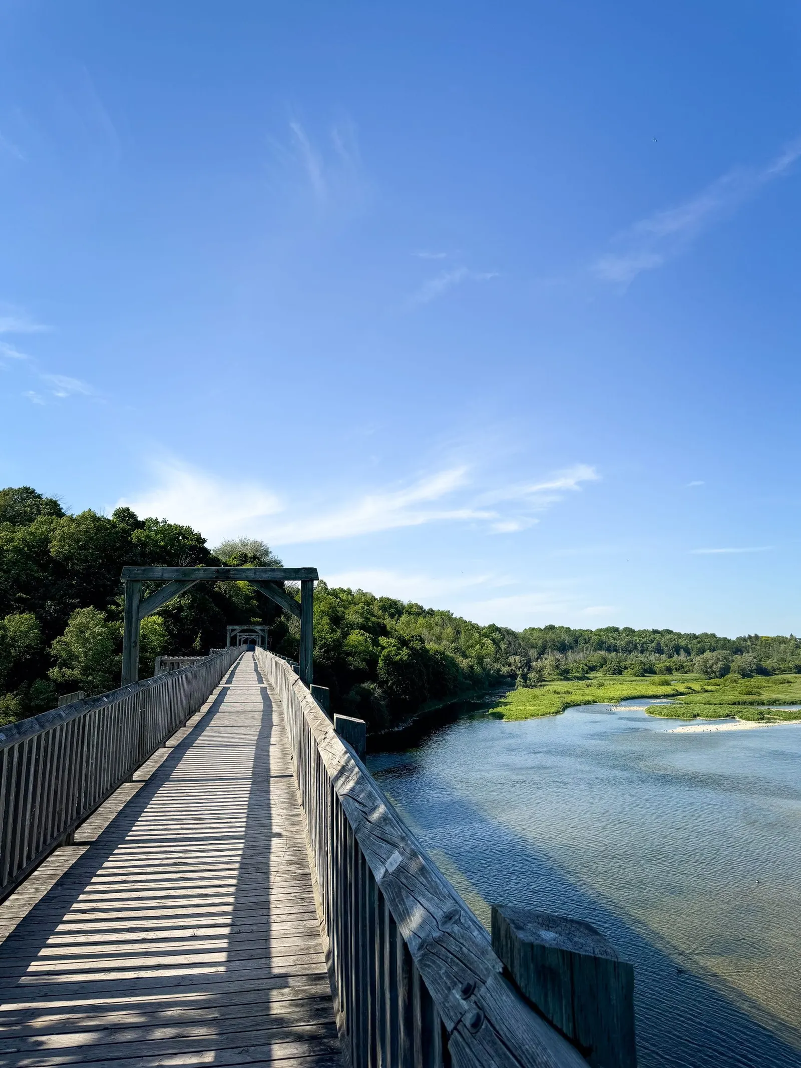 A long wooden rail bridge crossing a wide river, its boardwalk stretching into the trees