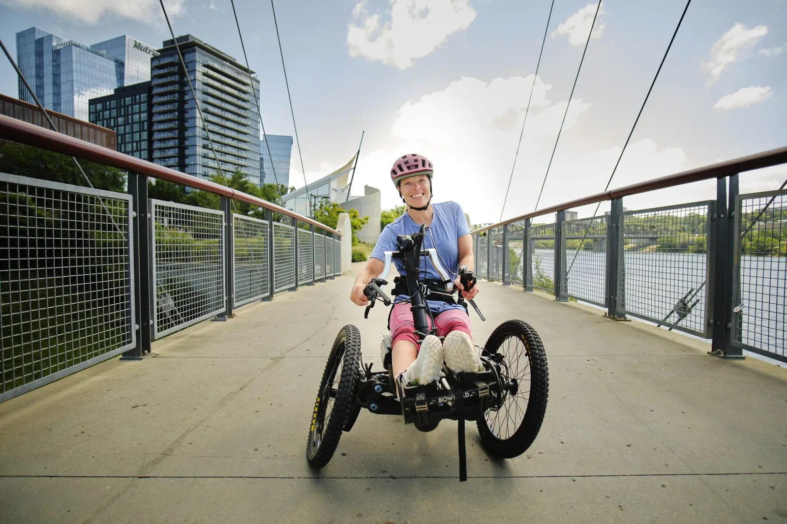 A smiling rider on an adaptive hand-cycle crossing a river bridge with the city skyline behind