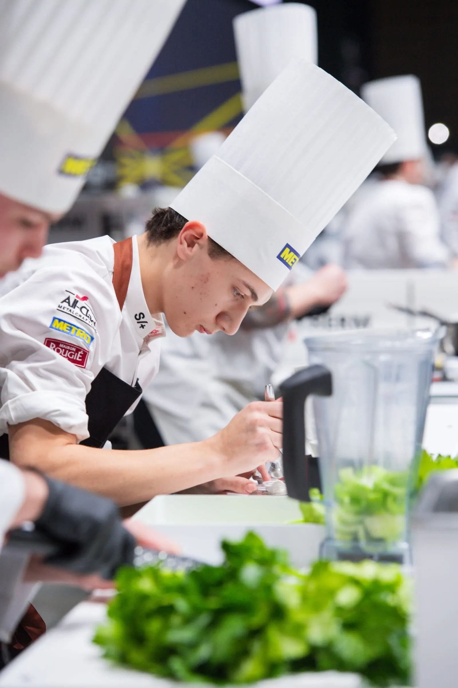 Commis Joseph Mongillo in his toque, head down over precision prep work beside a blender of greens at the competition bench