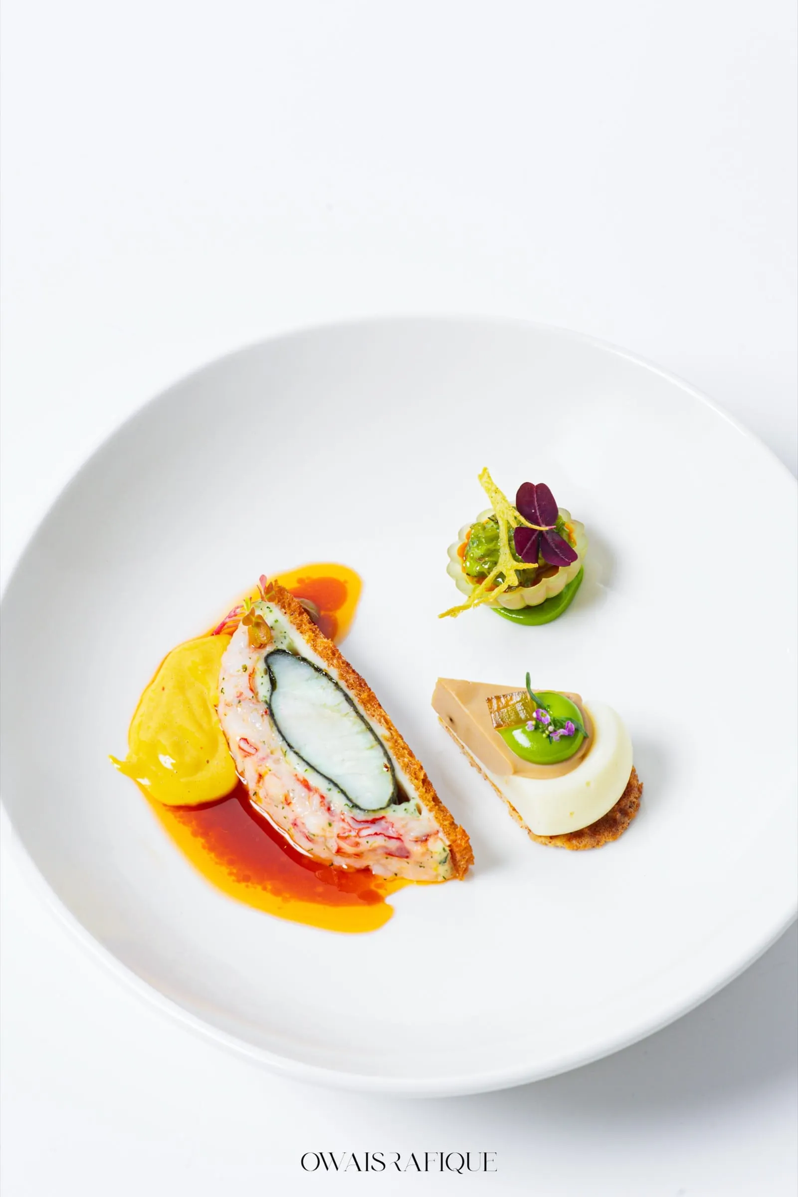 A composed competition plate: a crusted stone bass and lobster preparation beside a quenelle of yellow purée and two garnished bites, on a white plate
