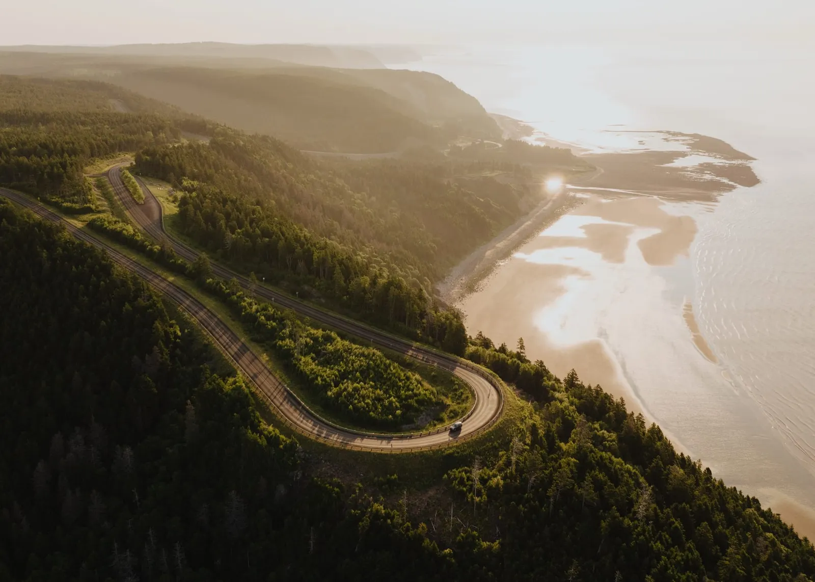 Aerial view of the Fundy Trail parkway winding through coastal forest above a sweeping beach at golden hour