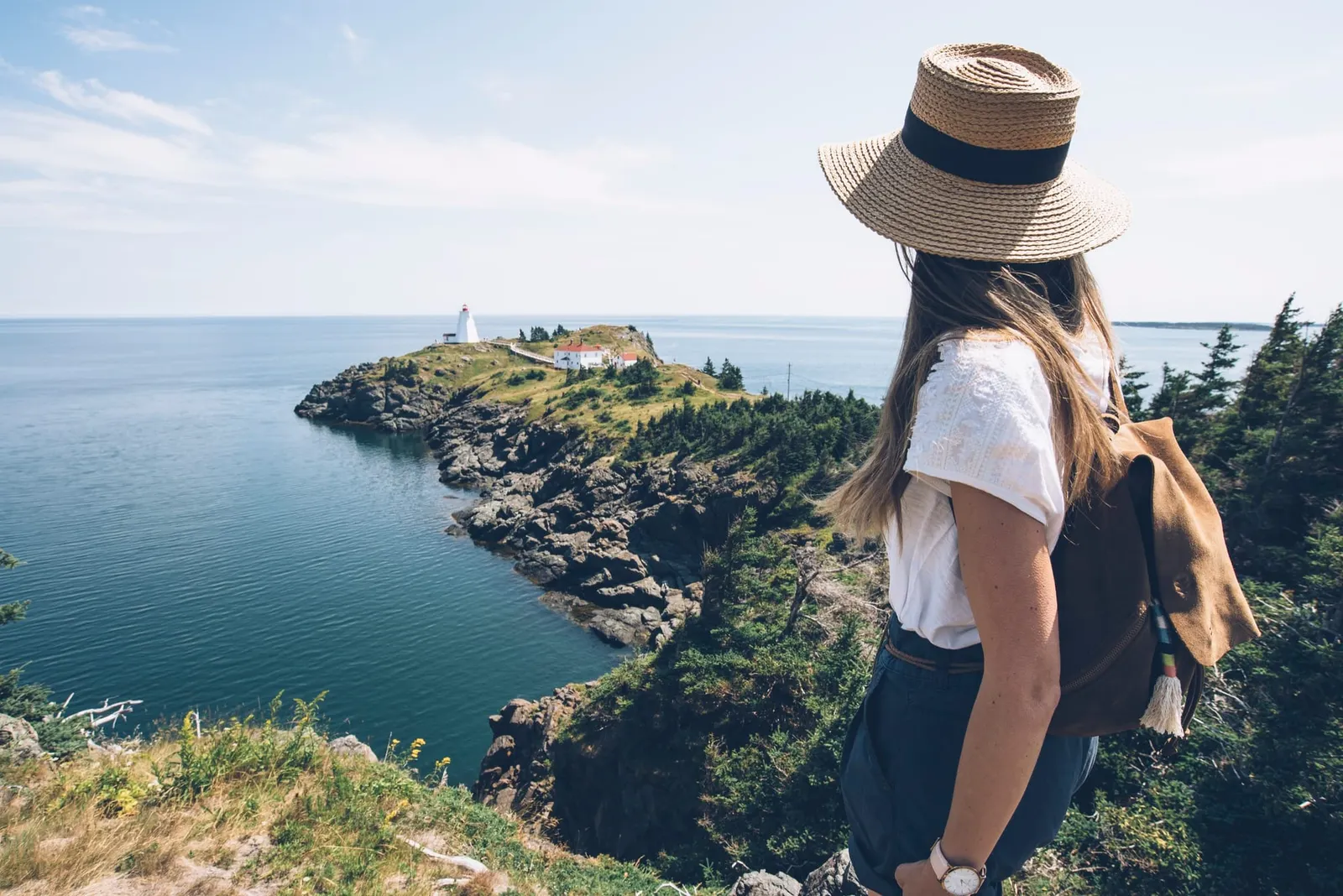 A traveller in a sun hat looking across a rocky cove toward a lighthouse on a headland
