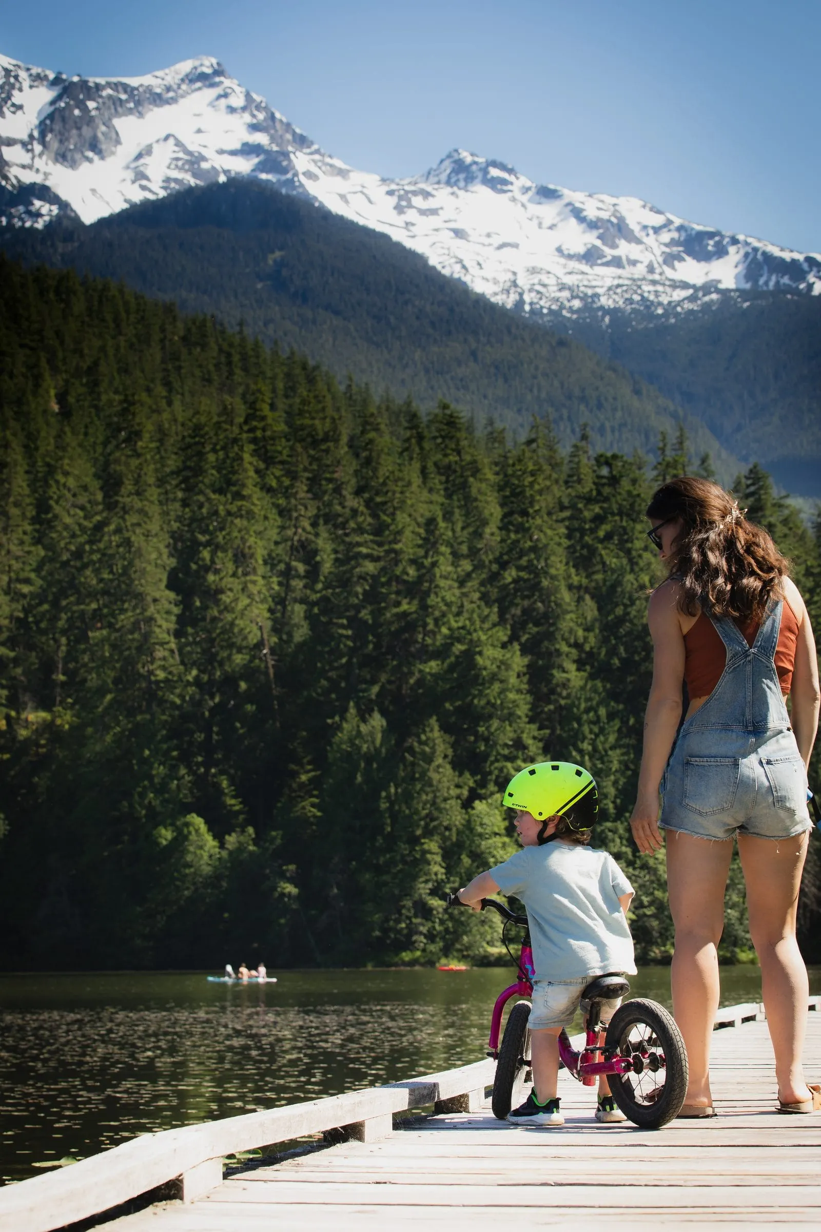 A parent and a child on a balance bike on a lakeside dock beneath snow-capped mountains