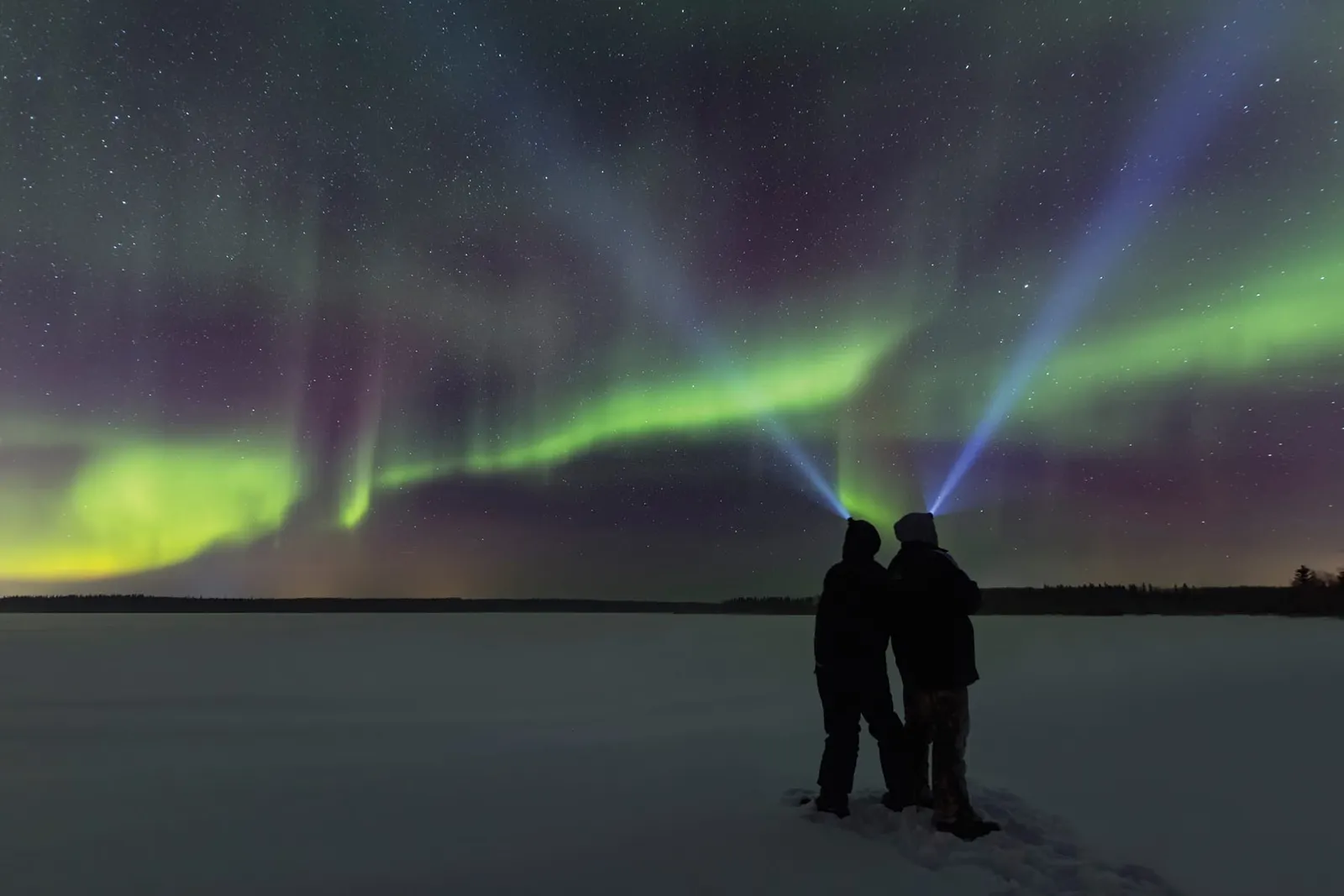 Two people with headlamps stand on a frozen lake under green and violet northern lights