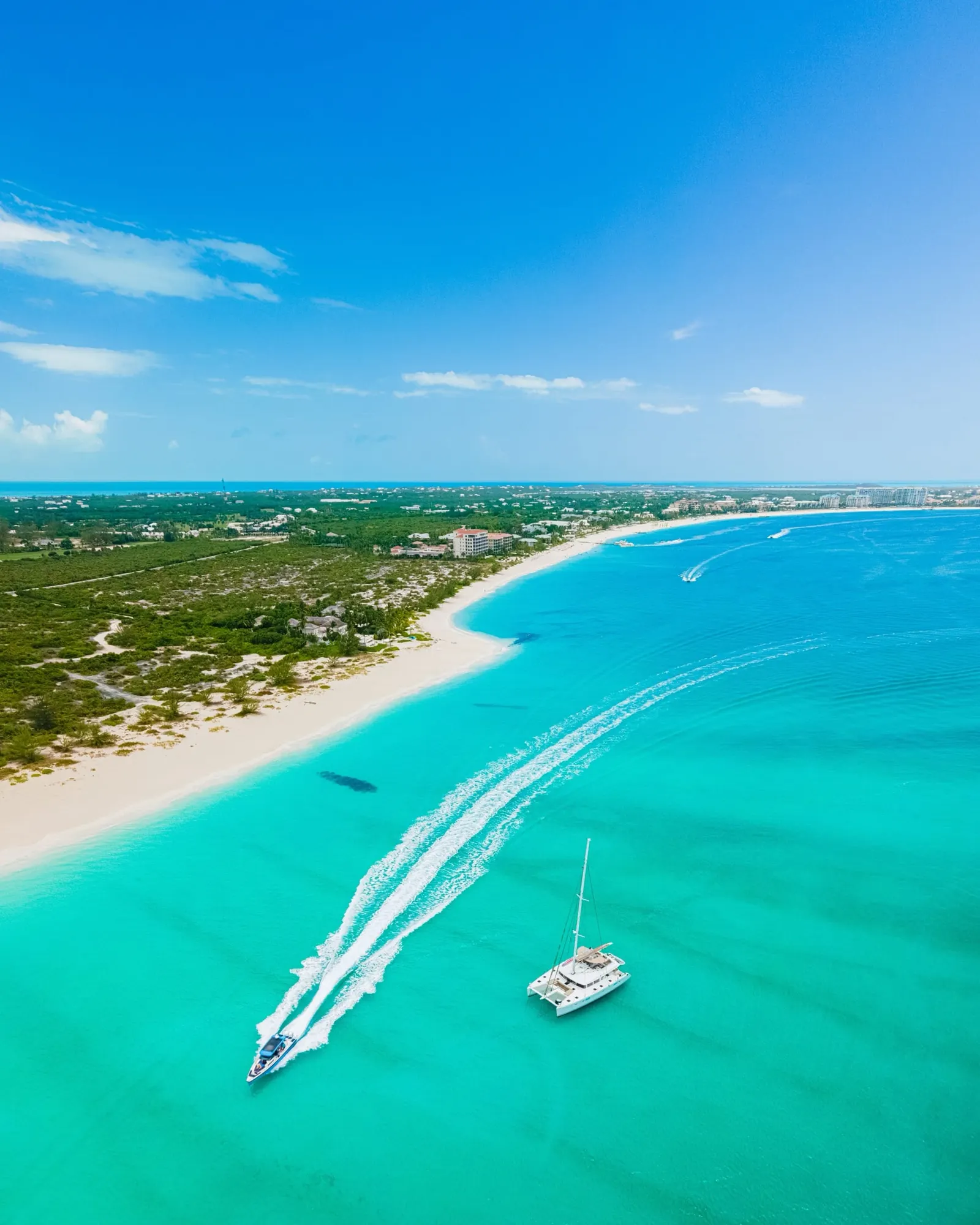 Aerial view of a long white beach and turquoise shallows with a catamaran and a boat wake