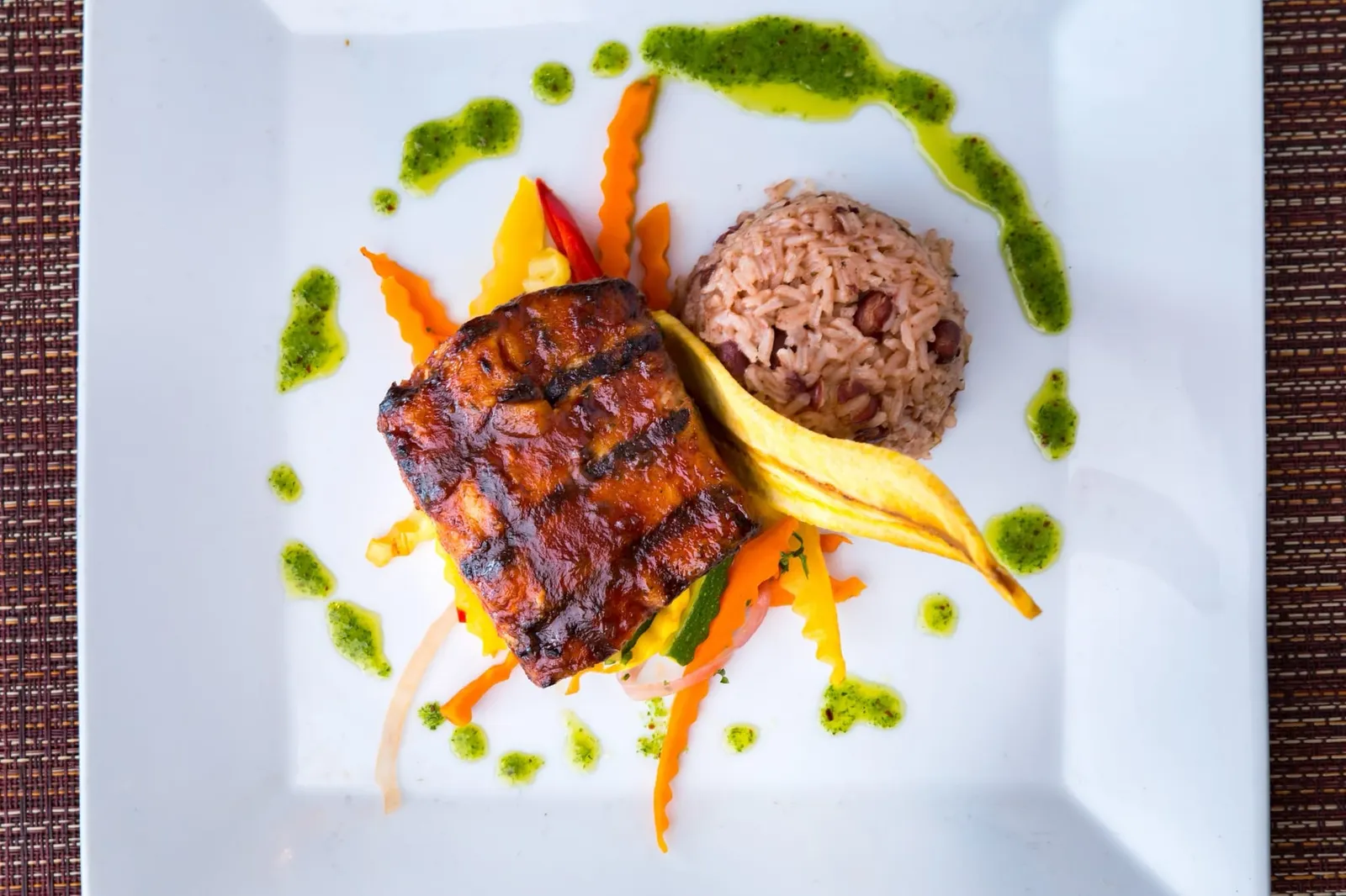 A grilled fish fillet with rice and peas, plantain and vegetables on a white plate