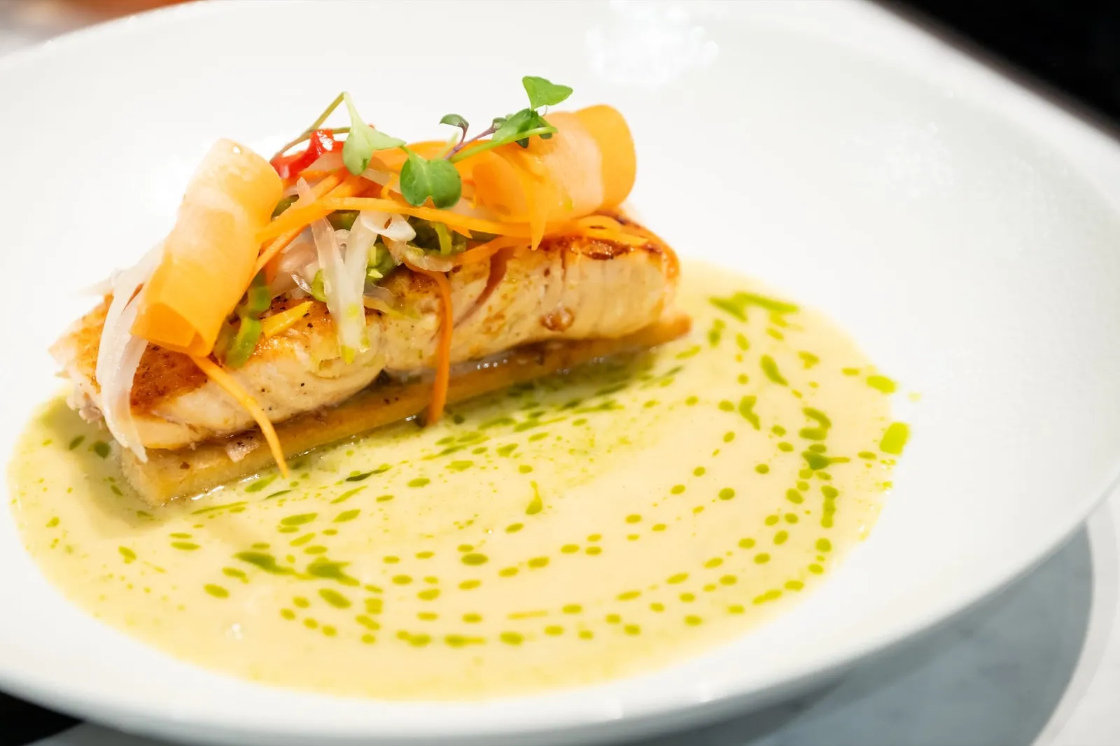 A fillet of fish topped with pickled vegetables on a pale sauce dotted with herb oil