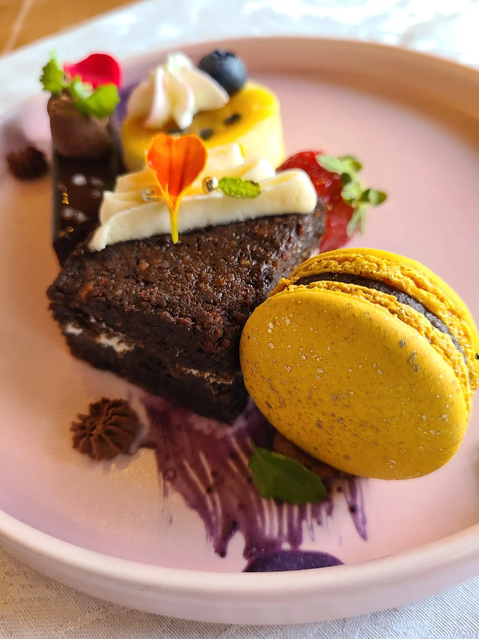 A slice of dark chocolate cake with a yellow macaron, fresh fruit and edible flowers