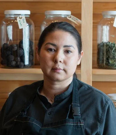 Chef Samantha Chevrier in a dark chef's jacket and apron, in front of wooden shelves of labelled glass jars of dried and preserved ingredients