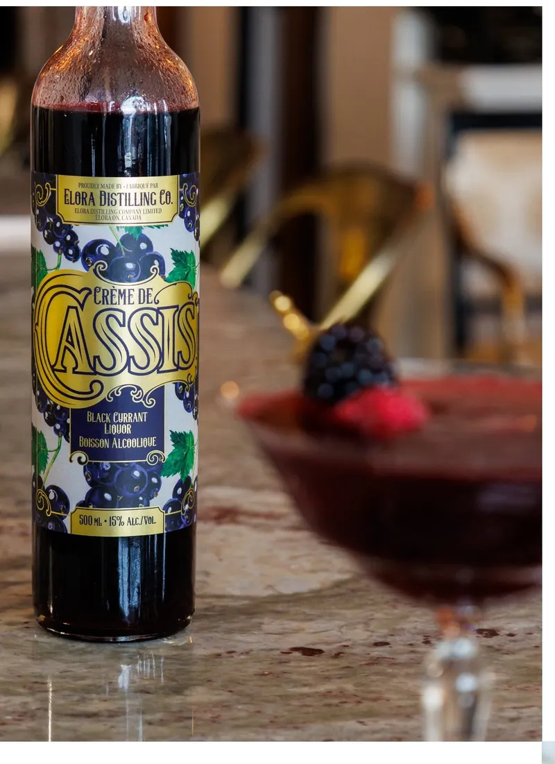 A bottle of Elora Distilling Co. Crème de Cassis beside a cocktail garnished with blackberries and a raspberry