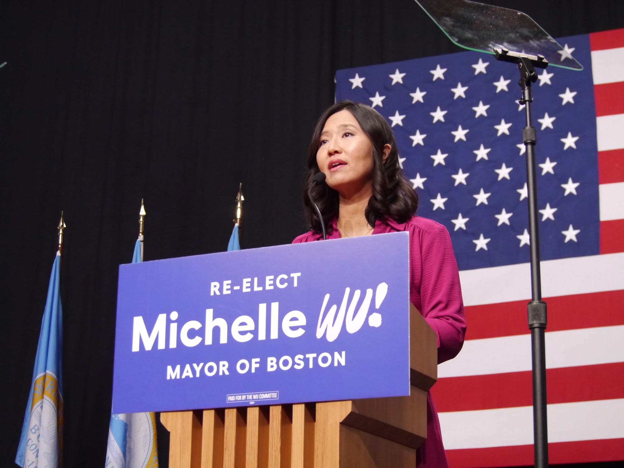 Boston Mayor Michelle Wu stands at a podium at the Boston Cyclorama with an American flag as a backdrop.