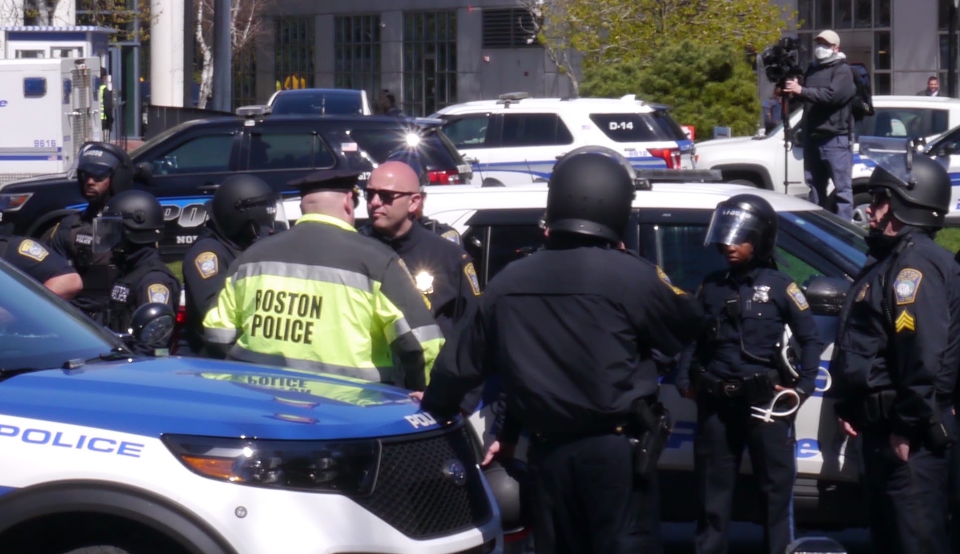 Boston police officers gather at the site of a Northeastern University student protest against Israel's siege of Gaza while a camera operator looms in background.