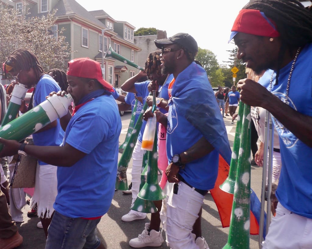 Members of Freda Rara march with large trumpet-like horns.