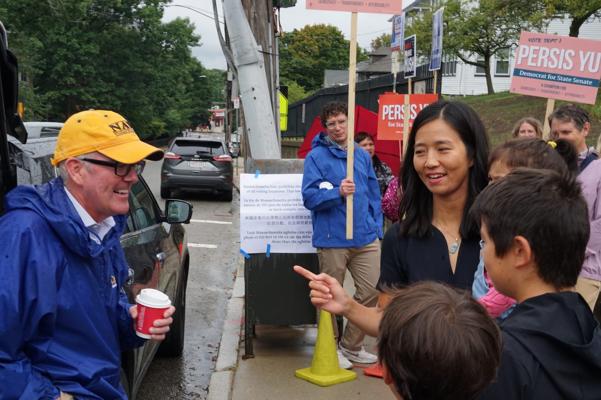 Mayor Michelle Wu gestures toward Sen. Michael Rush outside the Phineas Bates School in Roslindale while people holding Persis Yu signs look on..