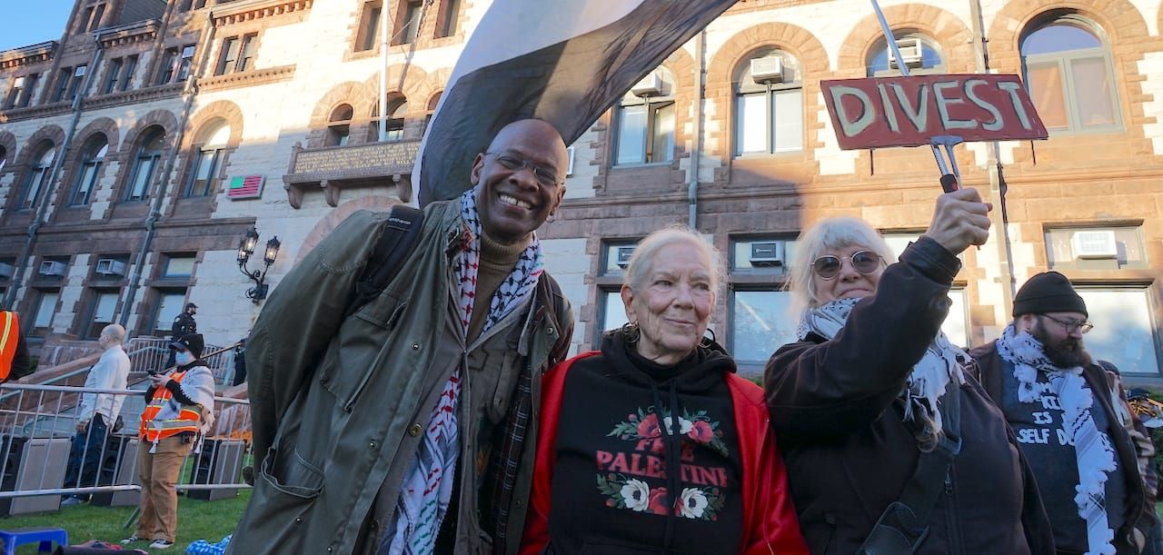 Ann Antonellis holds a sign that reads "divest", attached to a Palestinian flag, standing next to Michel DeGraf and Susan Escovitz in front of Cambridge City Hall.