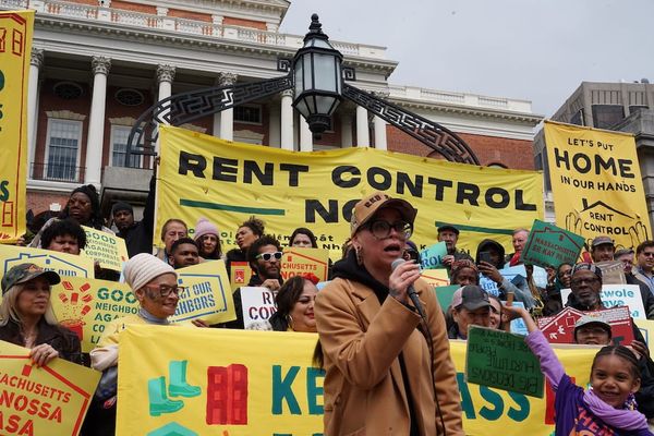 Mimi Ramos, who heads the New England Community Project, speaks on a microphone in front of rent control supporters holding signs in front of the State House Tuesday.