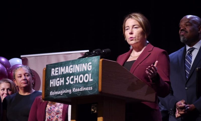 Governor Maura Healey speaks from a podium at Dedham High School as Education Secretary Paul Tutwiler and others look on.